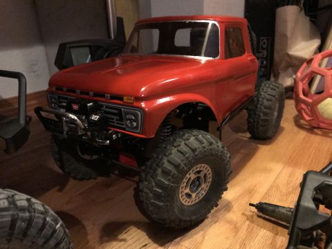 1989 Ford Bronco - A bright red, modified RC truck with large off-road tires and a winch mounted on the front bumper sits on a wooden floor. The vehicle appea…