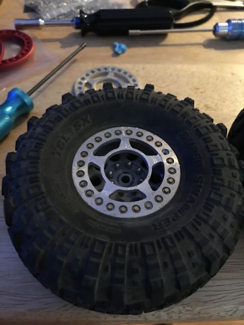 1989 Ford Bronco - A close-up view of a large, knobby RC car tire with a silver beadlock wheel. Tools and other RC car parts are visible in the background on…