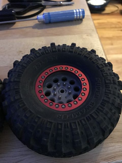 1989 Ford Bronco - A close-up view of a black RC car tire with a red beadlock ring and a gray wheel. The tire has "TSL SX" and "SUPER SWAMPER" text visible on…