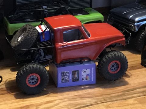 1989 Ford Bronco - A red RC rock crawler truck is shown from a 3/4 front angle, resting on a box. The truck features large off-road tires and a detailed inter…
