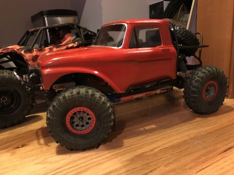 1989 Ford Bronco - A red RC truck with a classic pickup body style is parked on a wooden floor next to another RC vehicle with a flame graphic. The red RC tru…