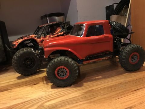 1989 Ford Bronco - Two remote-controlled rock crawlers are shown on a wooden floor. The one in the foreground is red with a roll cage and large tires, while t…