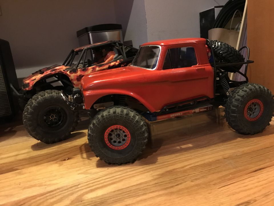 Two RC rock crawlers, one red and one with flame graphics, on a wooden floor.