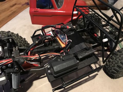 1989 Ford Bronco - A close-up view of the chassis and drivetrain of a red RC vehicle, possibly a rock crawler, showing suspension, wiring, and a black roll ca…