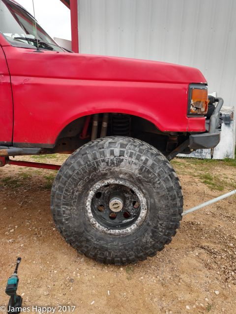 1989 Ford Bronco - A red Ford Bronco's front fender and large off-road tire are shown in this side view. A teal cordless impact wrench rests on the dirt groun…