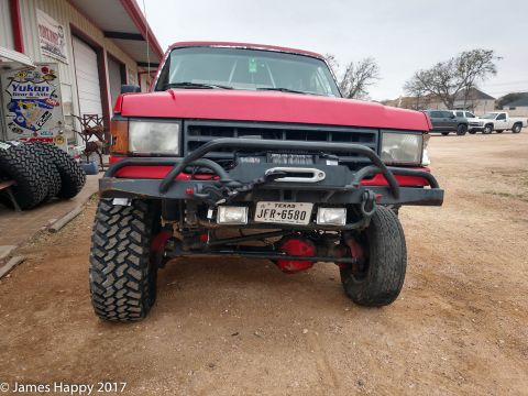 1989 Ford Bronco - Front view of a lifted red Ford Bronco with large off-road tires and a winch mounted on the aftermarket bumper. The vehicle is parked on a…