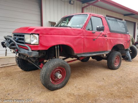 1989 Ford Bronco - A red, lifted Ford Bronco with a soft top is parked on a gravel surface in front of a building with large garage doors. The vehicle feature…