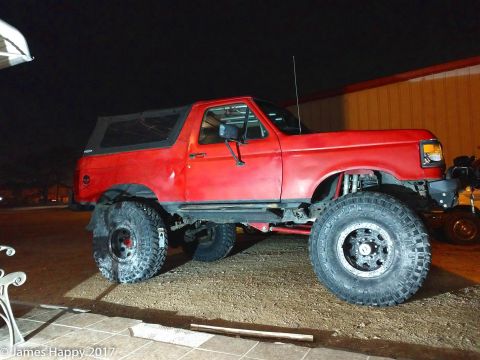 1989 Ford Bronco - A red, lifted Ford Bronco with large off-road tires is parked on a gravel surface at night. The vehicle has a soft top and a skull decal on…