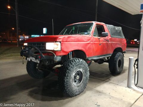 1989 Ford Bronco - A red Ford Bronco with large off-road tires and a black soft top is parked at a gas station at night. The vehicle features a custom front b…