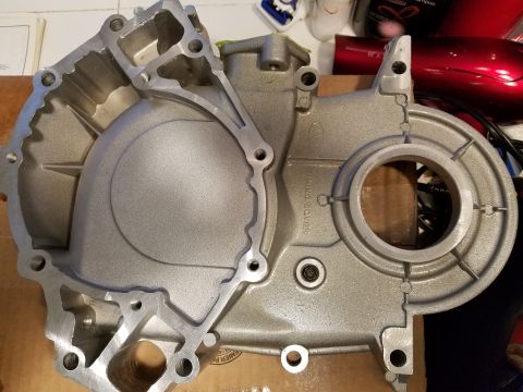 1976 Ford Ranchero - A silver aluminum timing cover is shown from a top-down perspective, resting on a cardboard surface. The cover has various bolt holes and a…