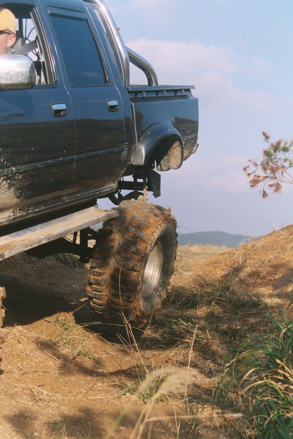 Lifted dark blue crew-cab pickup on huge muddy off-road tires climbing a dirt embankment