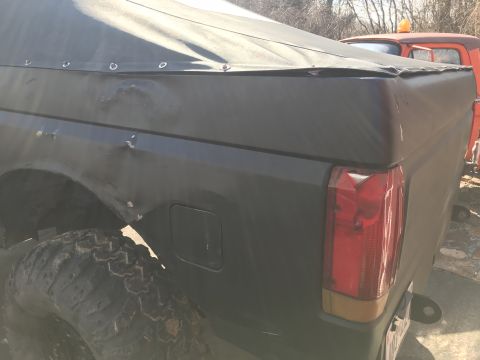 1989 Ford Bronco - The rear quarter panel and tailgate of a dark-colored vehicle are visible, showing significant body damage and a soft tonneau cover. A larg…