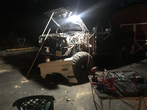 1989 Ford Bronco - A dark colored Ford Bronco is parked outside at night with its hood open, revealing the engine bay. Tools and power tools are laid out on a…