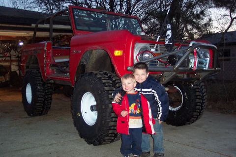 1974 Ford Bronco - A red 1974 Ford Bronco, modified with large off-road tires and a roll cage, is pictured with two young boys posing in front of it.