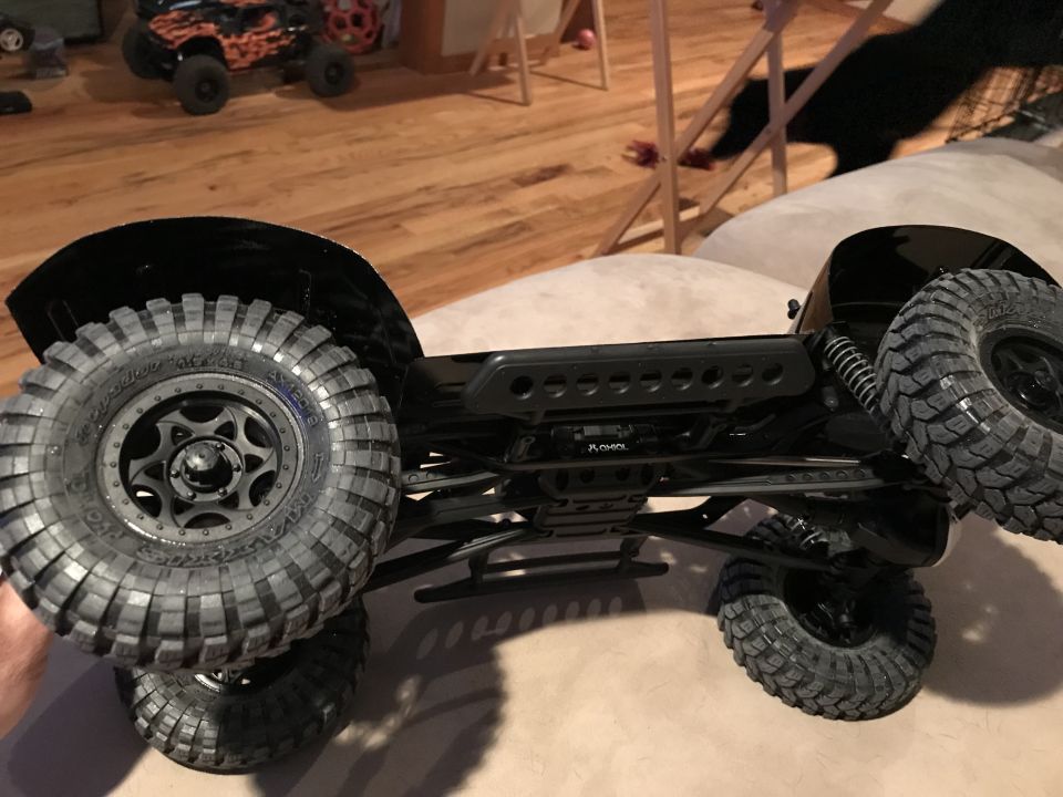 Black RC rock crawler underside with large tires and suspension, resting on a couch.