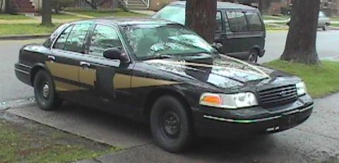 1998 Ford Crown Victoria - A black Ford Crown Victoria with a distinctive gold stripe is parked on a residential street next to a tree.
