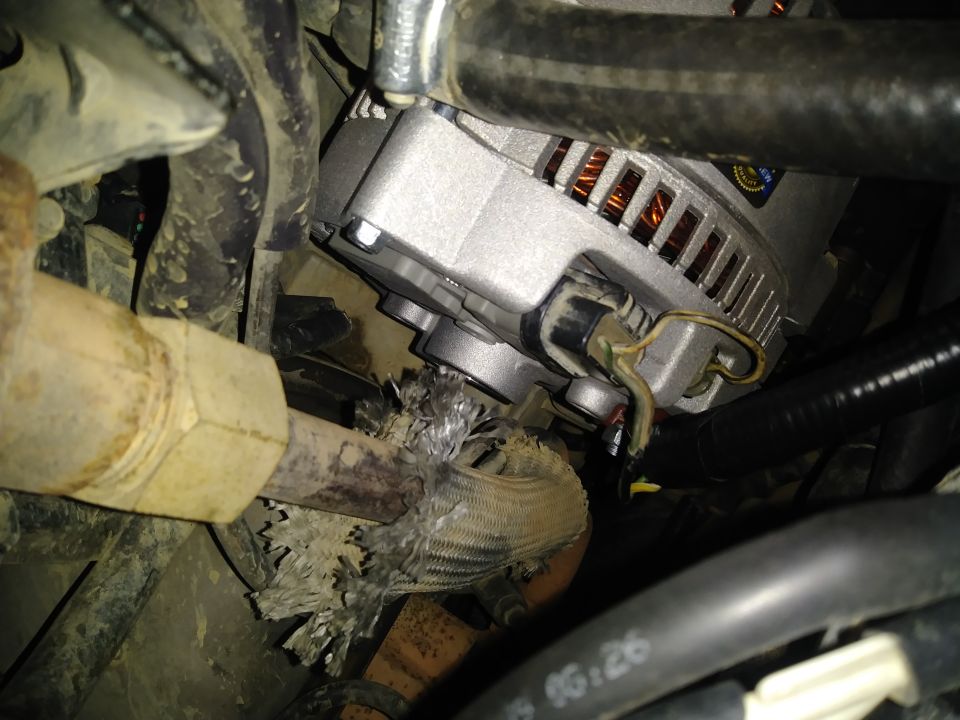 Close-up of a vehicle alternator with frayed wiring and a damaged exhaust pipe section