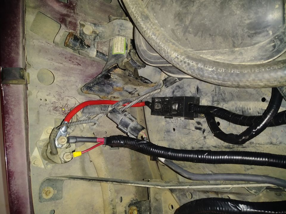 Engine bay wiring with red and black cables, a black fuse box, and a ribbed hose. The area is dirty and shows signs of rust