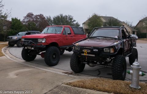 1989 Ford Bronco - Two modified trucks, a red Bronco and a maroon pickup, are parked on a residential street under an overcast sky.