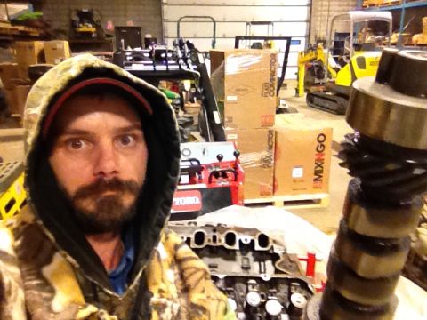 1979 Ford Bronco - A man takes a selfie in a workshop filled with engine parts and construction equipment, including a yellow mini-excavator.