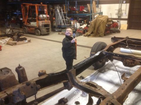 1979 Ford Bronco - A young child stands in a workshop, holding a paintbrush near a vehicle frame undergoing restoration.
