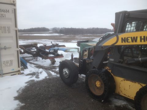 1979 Ford Bronco - A trailer carrying a vehicle chassis and parts sits in a snowy outdoor area next to a New Holland skid steer loader.