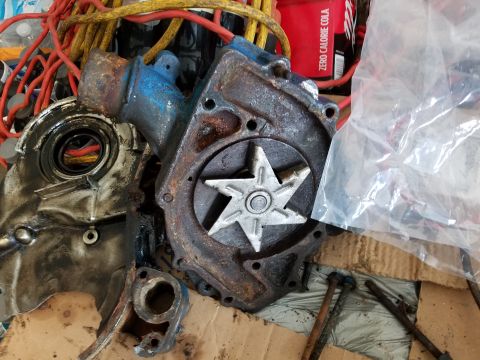 1976 Ford Ranchero - A rusty automotive water pump and timing cover are shown with various wires and a can of Coca-Cola in the background.