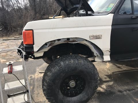 1989 Ford Bronco - A white Ford F-250 with a damaged front fender and a large off-road tire is shown, with the hood open and tools nearby.