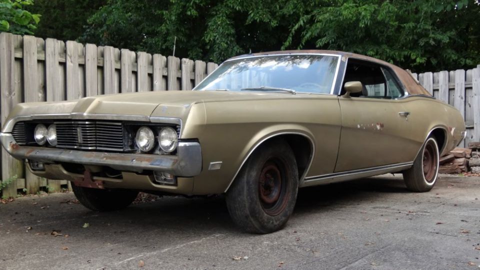 Gold 1969 Mercury Cougar coupe with a rusty front bumper and aftermarket wheels parked outdoors