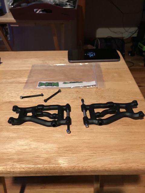 1989 Ford Bronco - Two black plastic suspension control arms and hardware are laid out on a wooden table next to a phone and a sticker.