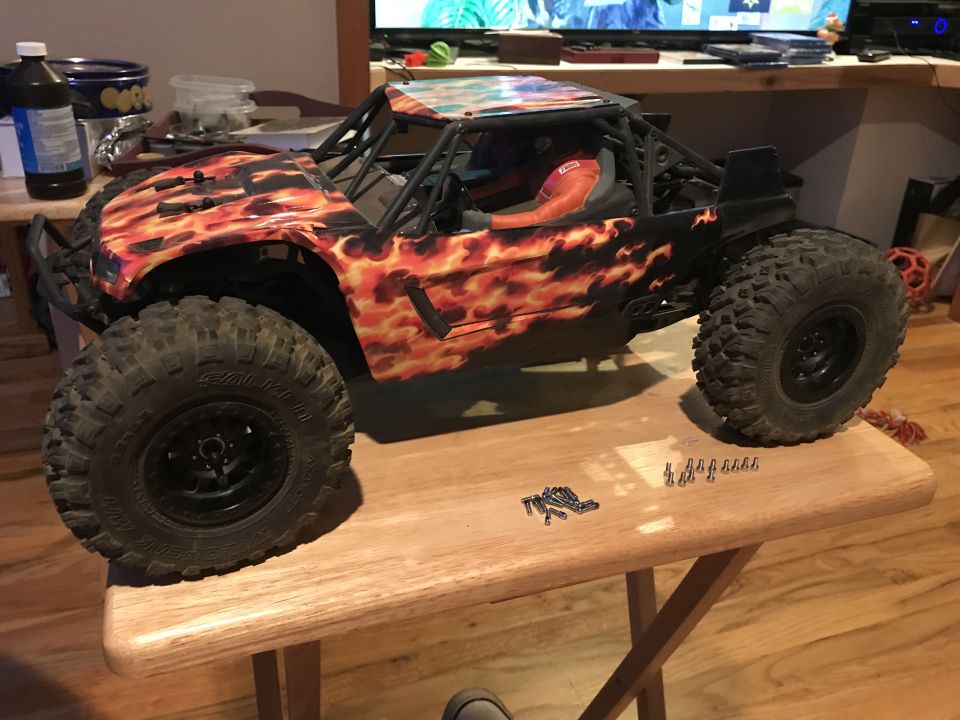 Remote control rock crawler with flame graphics, black roll cage, and large off-road tires, sitting on a wooden table