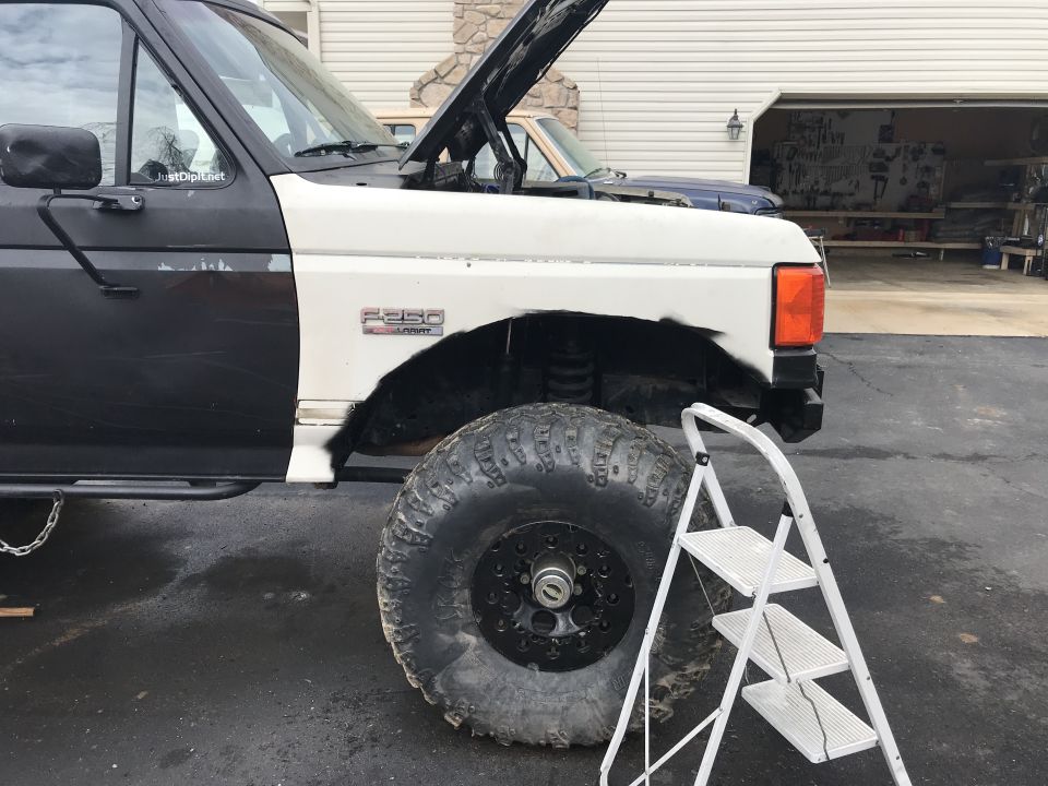 Black and white Ford F-250 truck, hood open, large off-road tire, ladder nearby