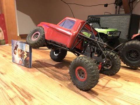 1989 Ford Bronco - A red remote control rock crawler truck is positioned on a wooden floor next to a DVD case.