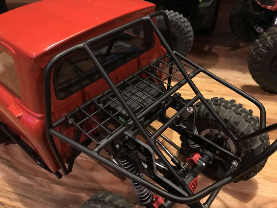 Red RC rock crawler truck with a black roll cage and spare tire mount. Other RC vehicles are visible in the background