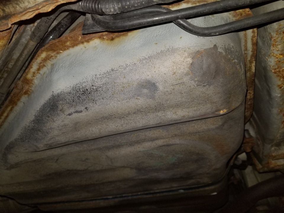 Underside view of a vehicle's fuel tank and rusted frame components. Some fuel lines are visible above
