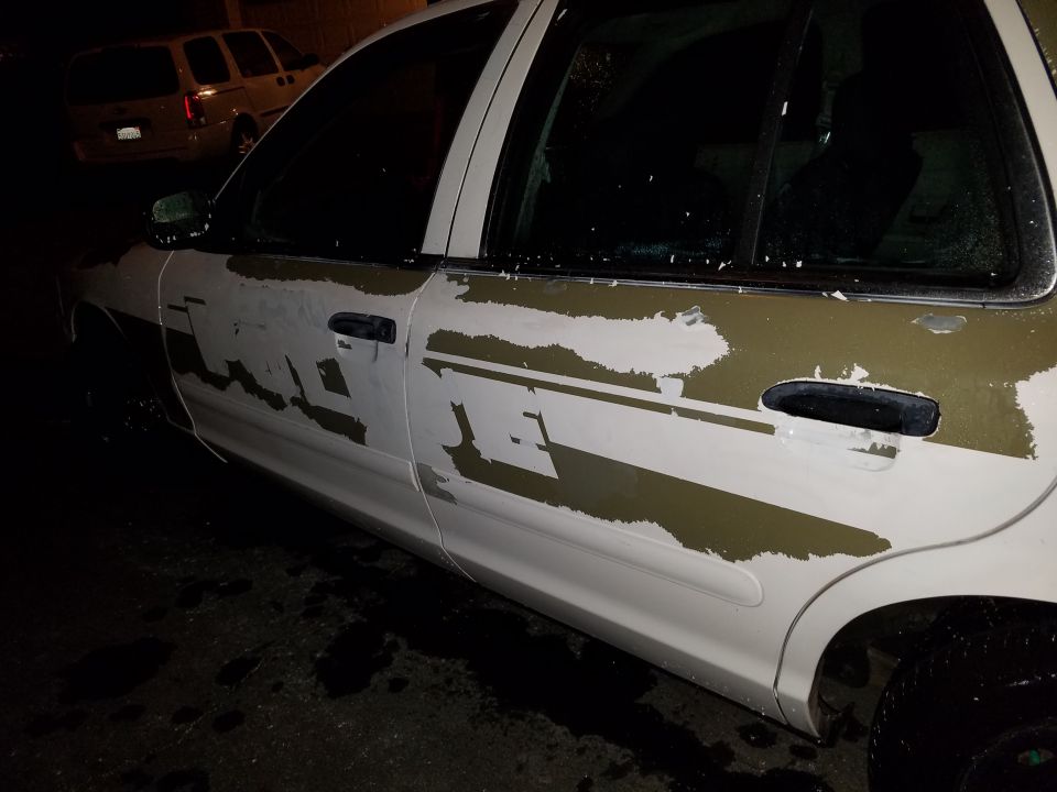 Side view of a white Ford Crown Victoria with peeling green and white decals. Another SUV is parked behind it