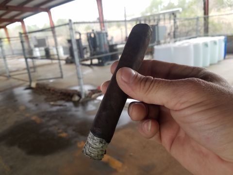 2018 Ford Bronco - A hand holds a dark brown cigar with a white band at the end. The background shows an outdoor industrial area with a chain-link fence and l…