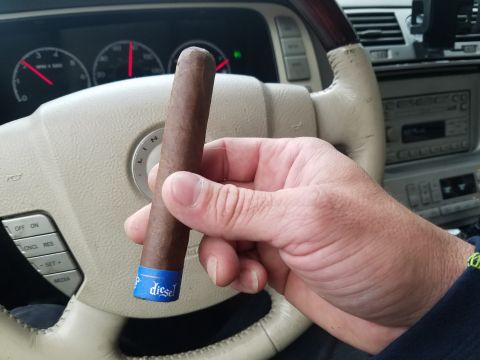 2018 Ford Bronco - A hand holds a Diesel brand cigar in front of a car's steering wheel and dashboard. The steering wheel has controls for cruise control and…