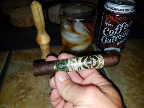 2018 Ford Bronco - A hand holds a Perdomo cigar in front of a glass of amber liquid with ice and a can of "Coffee Oatmeal Stout". A wooden cigar press is visi…