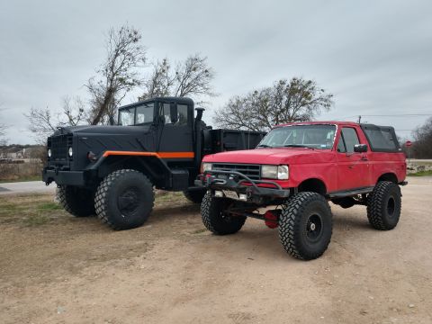 1989 Ford Bronco - A lifted red 1989 Ford Bronco is parked next to a lifted black military-style truck on a dirt lot under a cloudy sky.