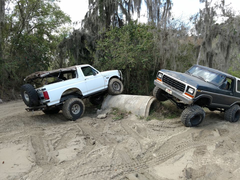 Two Ford Broncos off-roading, one white and one dark blue, over rocks and a concrete pipe.