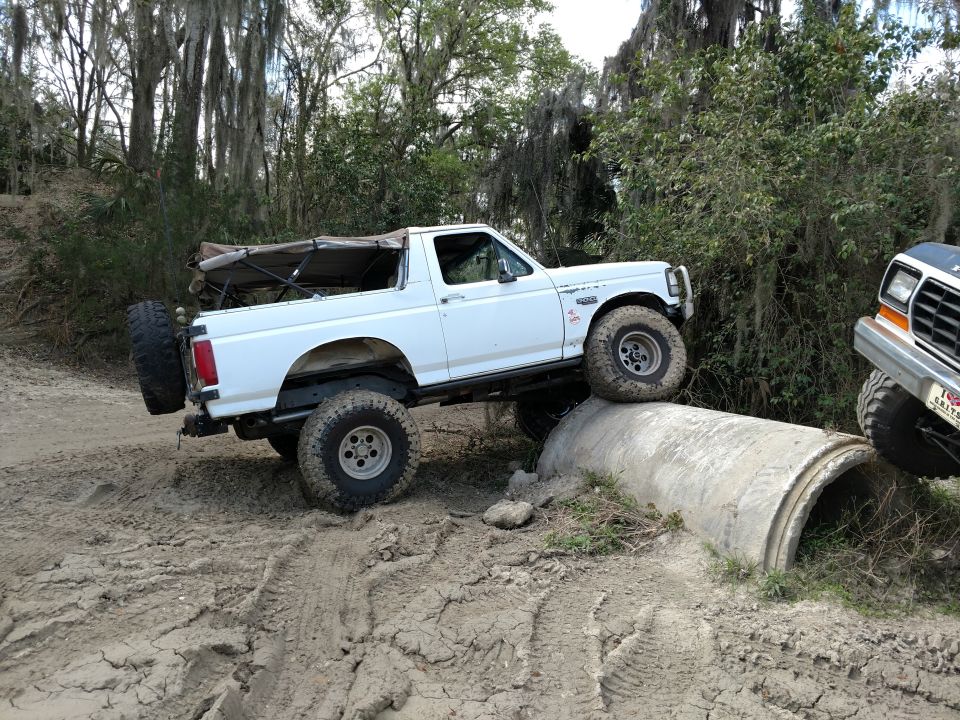 White Ford Bronco driving over a concrete pipe on a muddy trail.