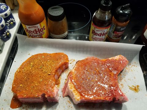 2018 Ford Bronco - Two raw pork chops seasoned and covered in sauce are laid out on parchment paper, with bottles of Frank's RedHot sauce and various spice sh…