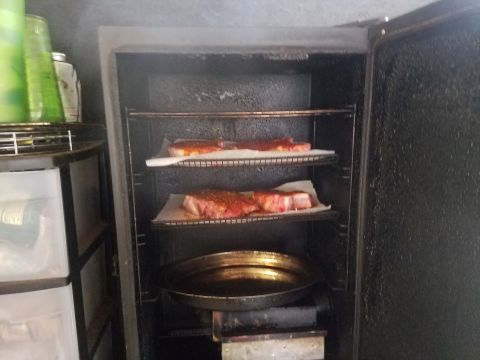 2018 Ford Bronco - Two raw steaks seasoned with spices are placed on racks inside a smoker. Below the racks, a metal pan sits on a heating element.