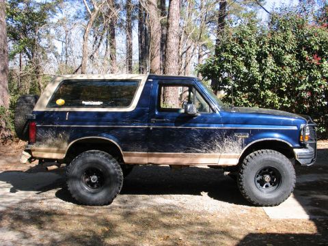 1995 Ford Bronco - This blue Ford Bronco has a tan removable top and is lifted with large off-road tires. Mud covers the lower body panels.