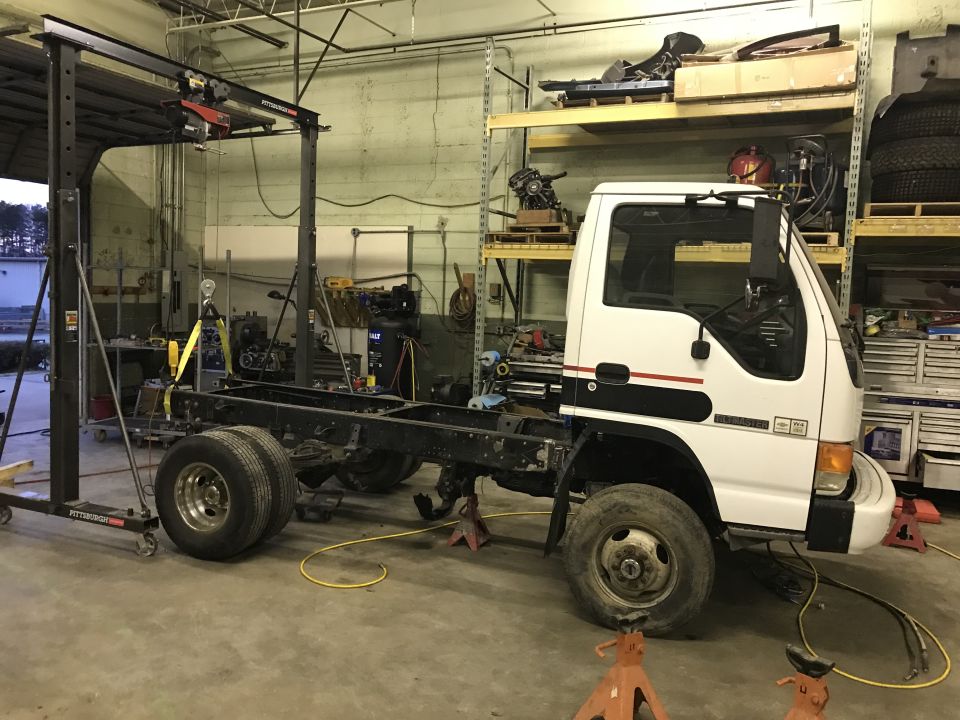 White truck chassis on jack stands with engine hoist overhead in a workshop.