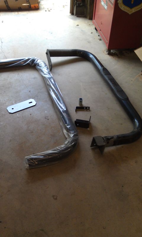 1995 Ford Bronco - Two black metal nerf bars or side steps are laid out on a concrete floor, along with several mounting brackets and hardware. One of the bar…