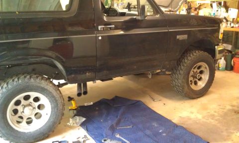 1995 Ford Bronco - The side view of a black Ford Bronco is shown in a garage setting. The vehicle has large off-road tires and dual exhaust pipes visible.