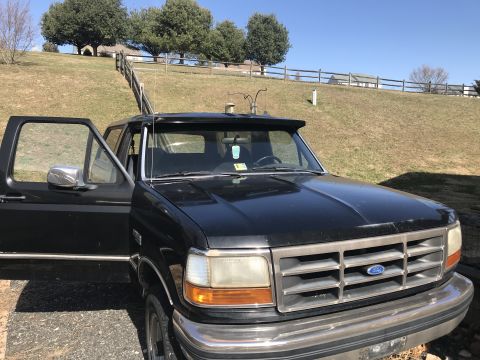 1992 Ford Bronco - This black Ford Bronco is parked on a paved surface with a grassy hill and fence in the background.