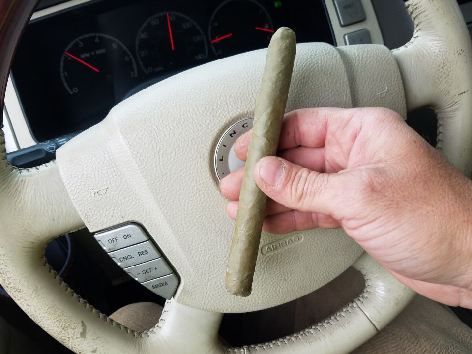 Hand holding a cigar over a Lincoln steering wheel with dashboard gauges visible